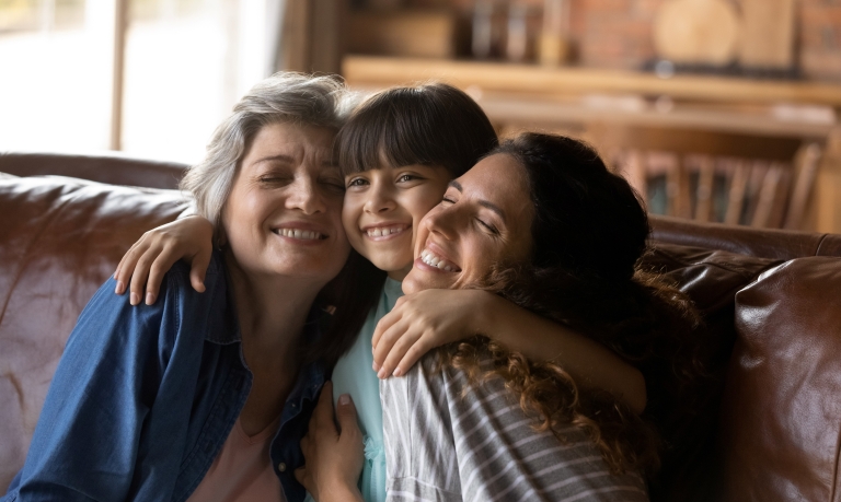 Child hugging her grandmother and mother while sitting with both on a leather sofa.