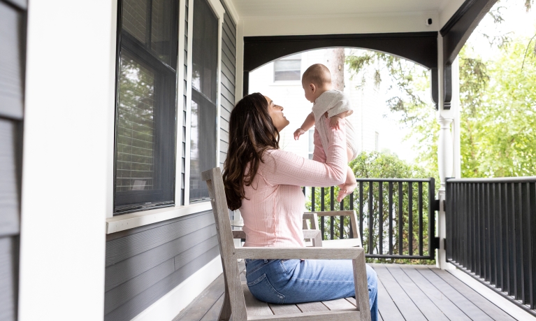 Mom sitting on a rocking chair on the porch of her house while holding her baby up.