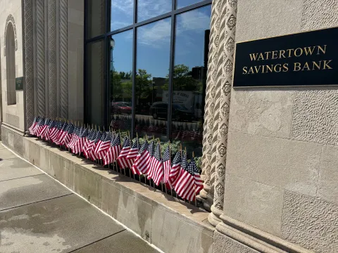American flags placed standing up alongside a wall of windows of the Watertown Savings Bank building.
