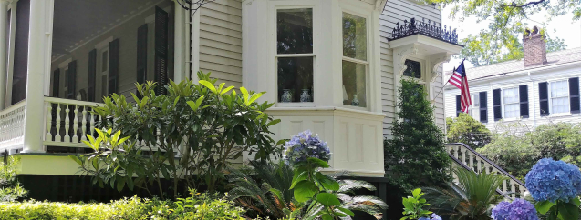 A colonial house with a porch and large window with beautiful green landscaping and hydrangeas in front.