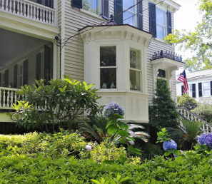 A colonial house with a porch and large window with beautiful green landscaping and hydrangeas in front.