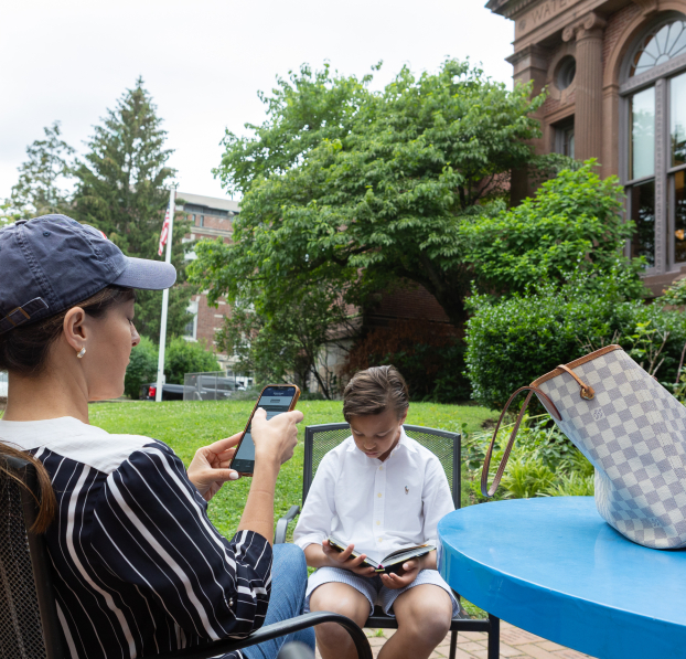 Mom checking her phone and son with a book sitting at a table outside of the library.